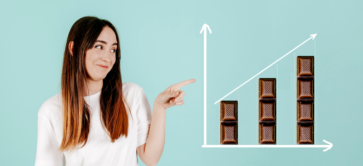 Grafika przedstawia dziewczynę wskazującą na wykres z trzema słupkami, które są zbudowane z kostek czekolady,